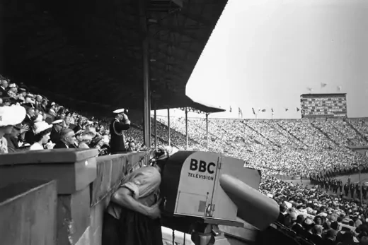 A BBC camera operator films the opening ceremony in a crowded stadium.