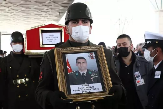 A Turkish officer in a face mask holds an image of Lieutenant Burak Coskun. Officers behind him carry Coskun's coffin.