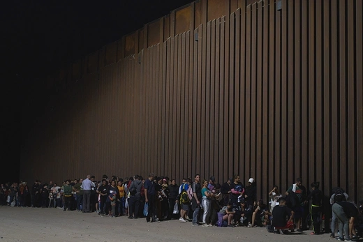 A line of immigrants waiting at night by the border wall between the U.S. and Mexico.