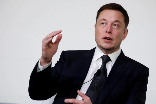 Elon musk looks towards the top left of the frame and raises his hand in front of an off white background.