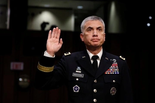 General Paul Nakasone stands in his dress uniform with his right hand raised in front of a dark background.