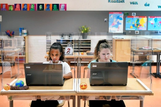 Two girls, one wearing a white shirt and another a blue shirt, sit behind plexiglass dividers with a laptop computer in front of them.