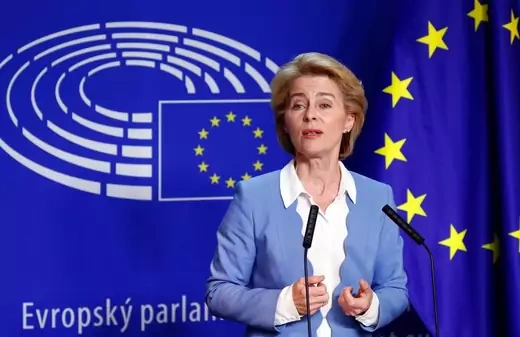 Ursula von der Leyen stands in front of a European Union flag, with two microphones in front of her.