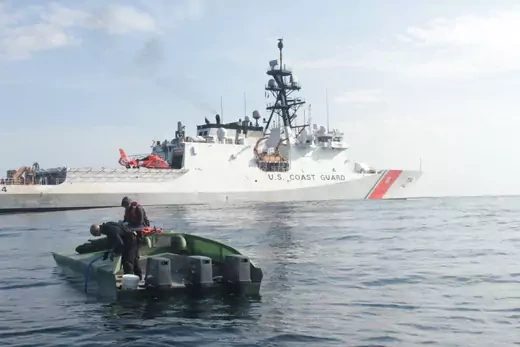 Two Coast Guardsmen search a low profile vessel in front of a large, white Coast Guard ship. 