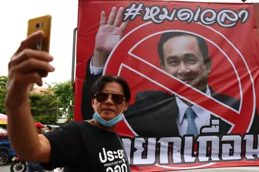 A protester in a black shirt takes a photo in front of a banner protesting the Thai prime minister