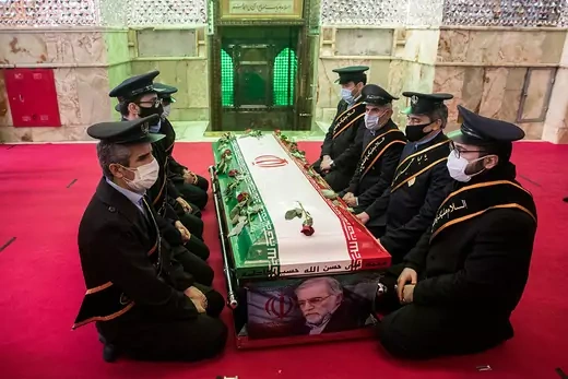 Men in military uniforms sit on either side of a coffin with Mohsen Fakhrizadeh's photo on one end of it