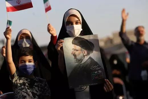 A young Iranian woman holds a portrait of Ebrahim Raisi. Other adults and a child are seen, some with small Iranian flags.