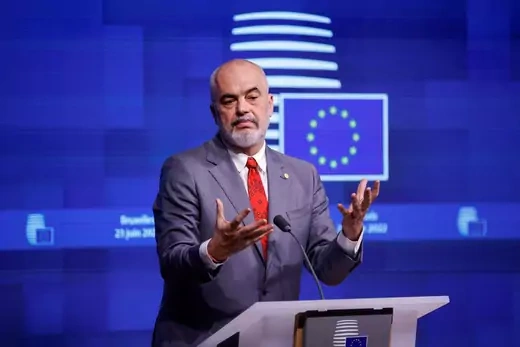 Edi Rama, dressed in a grey suit, stands in front of a lectern with his hands raised in front of his chest and a European Union flag projected in the background