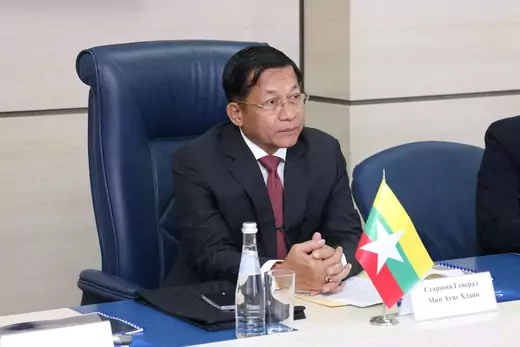 Man in suit sits at table with Myanmar flag in front of him