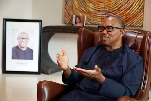 Peter Obi, a candidate for the upcoming Nigerian presidential election, sits on a brown chair and gestures. A framed photograph depicting himself hangs next to his brown chair.  
