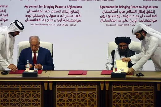 Zalmay Khalilzad sits at a table next to Abdul Ghani Baradar. Two other men hand them each a stack of documents.
