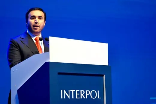 Ahmed Nasser Al-Raisi stands in front of a blue podium with Interpol written on it and a light blue background.