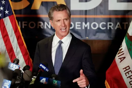 Gavin Newsom stands in front of a podium with a political sign in the background and an American flag on the left and California flag to the right.