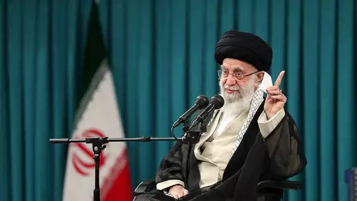 Iranian Supreme Leader Ali Khamenei gestures and speaks in to a microphone while seated. The Iranian flag is seen behind him.