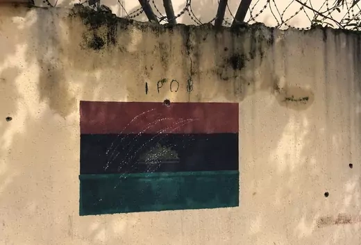 A flag is painted on a dirty wall with a barbed wire in the background. The flag is with a red, black, and green stripe. In the center of the flag, there is a half of a yellow sun rising.