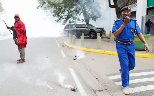 A man wearing red stands in the middle of the road filled with tear gas, while a police officer dressed in blue blows a whistle.