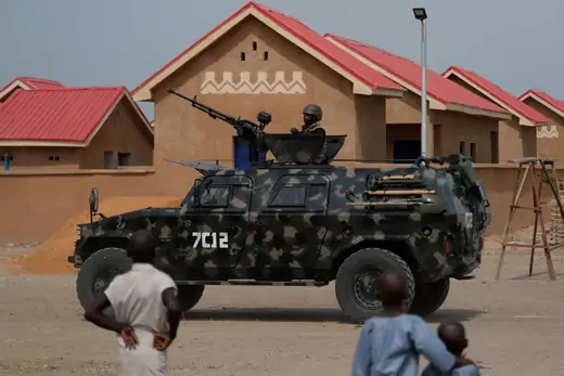 Nigerian armed forces point large rifles from an official army truck while Nigerians onlookers stand on the side of the dirt road.