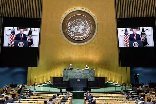 People sitting spaced apart at long tables watching a speech by Trump, which is shown on two screens that flank a podium at the UN headquarters.