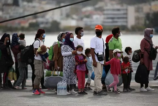People, including children, clump together with water and buildings behind them.  Some are wearing masks and carrying bags.