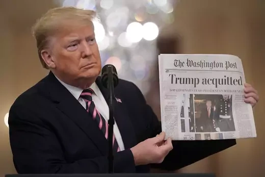 Dressed in a suit and standing in front of a microphone, Trump holds up a copy of the Washington Post showing news of his acquittal.