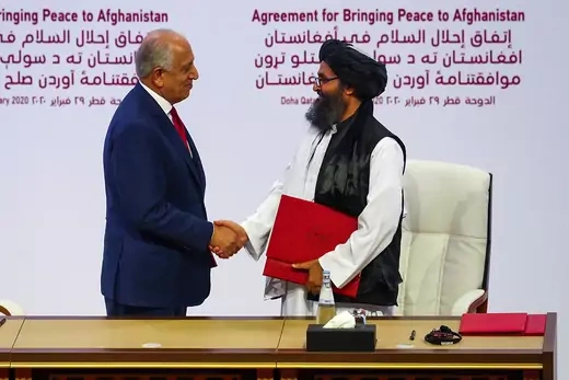 A photo of two men shaking hands in front of a screen that reads "Agreement for Bringing Peace to Afghanistan."  The man on the left is dressed in a navy-colored suit, and the man on the right has a beard, is wearing a head covering, and is carrying a red folder.