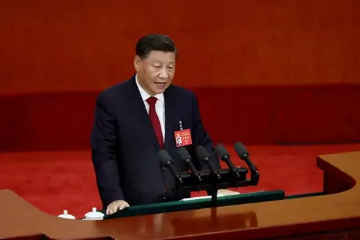 Chinese President Xi Jinping stands at speaker's podium in a black suit with a red tie, against a red backdrop.