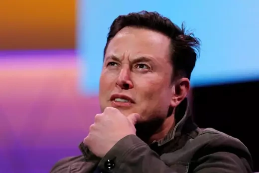 Elon Musk puts his hand on his chin, with a multi-colored screen in the background.