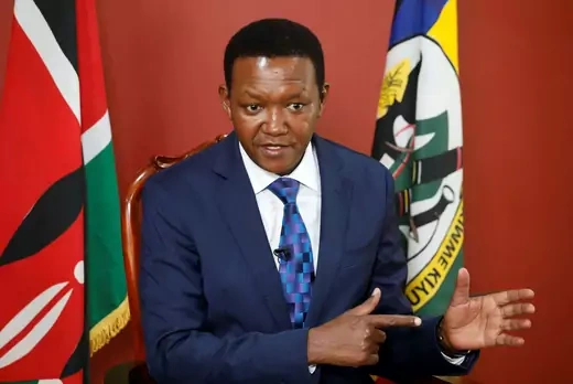 Dr. Alfred Mutua, Kenya's new Minister of Foreign and Diaspora Affairs, gives a speech in a blue suit, in front of a red background and a Kenyan flag. 