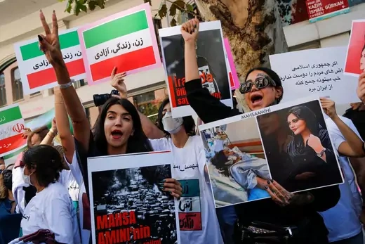 Protestors hold Iranian flags with slogans emblazoned on them and pictures of Mahsa Amini. Two women stand in the foreground with their fists raised, holding signs in their other hand.