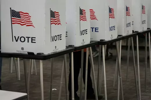 Picture shows tables with voting booths with the message, "vote" and an American flag. 