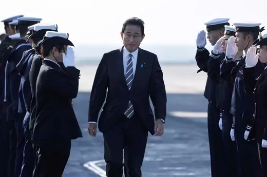 PM Kishida, in a dark suit, walks facing the camera between two rows of saluting soldiers.