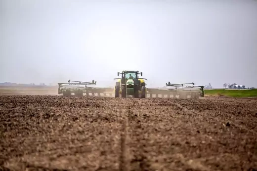 A tractor pulling a ripper sits in the middle of the frame, with green fields on the right third of the picture, and an empty field covering the rest.