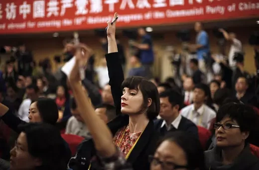 A female journalist raises her hand while sitting amongst a gathering of journalists.