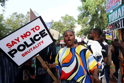 A woman wearing a colorful shirt holds a large sign that reads "Say No to Police Brutality" in a protest.