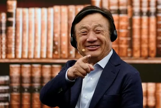 Ren Zhengfei stands in front of a bookshelf filled with brown volumes, and smiles while pointing his finger toward the camera.