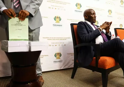 Sitting on an orange chair, beside a table with several stacks of paper covered in green film, South Africa's former Chief Justice awaits the handing over of the report on the Phala Phala scandal in Cape Town, South Africa.