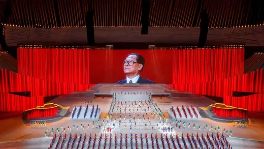 A photo of Jiang Zemin appears on a big screen as performers dance around it on a stage.