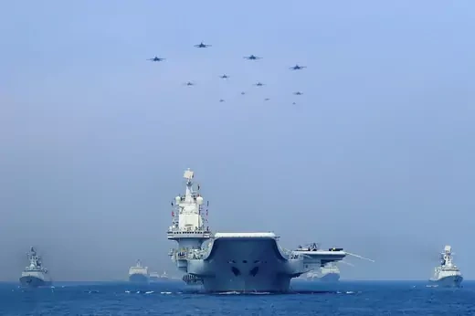 A Chinese aircraft carrier sits in the center of the screen, viewed even with the bow. It is flanked by five other smaller Chinese ships with eight aircraft directly overhead the carrier.