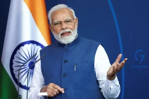 Narendra Modi stands in front of a blue background with his hand raised. An Indian flag hangs in the left of the frame.