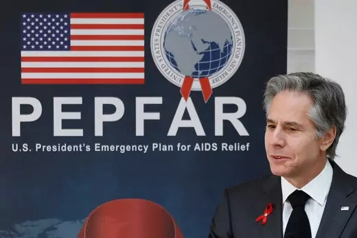 U.S. Secretary of State Antony Blinken, wearing a red ribbon pin for World Aids Day, stands in front of a blue sign reading 'PEPFAR U.S. President's Emergency Plan for AIDS Relief' in bold white letters. 