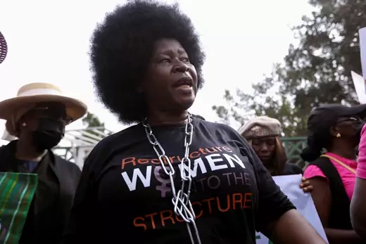 A woman wearing a "Restructure Women to the Structure" shirt protests alongside other women in Nigeria. She also wears a chain necklace around her neck. 