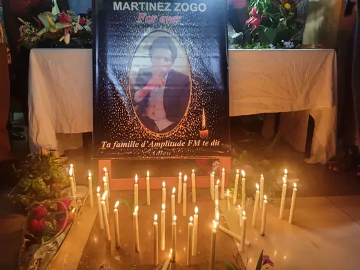 A vigil is shown with dozens of lit candles for prominent journalist Martinez Zogo. A poster of his face is shown which reads "Ta famille d'Amplitude FM te dit." 