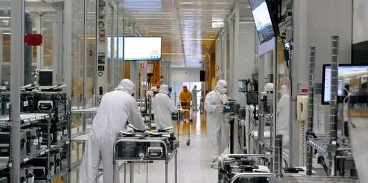 Workers wearing white PPE move stacks of semiconductors around a large room with glass walls.