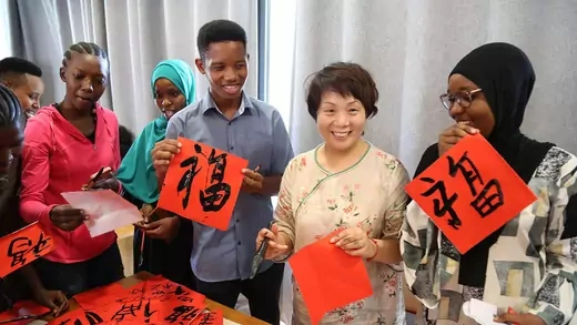 A woman stands beside students holding up their calligraphy painted on red paper.