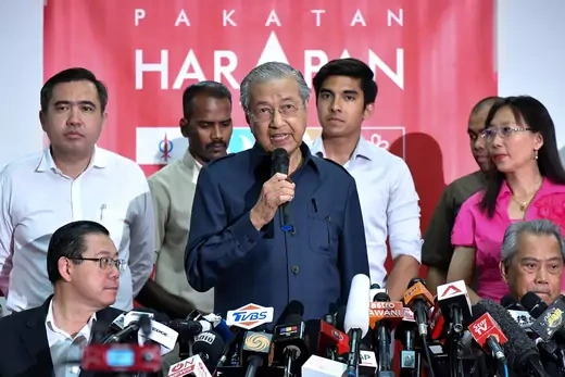 Newly elected prime minister of Malaysia stands in front of microphones with a red background.
