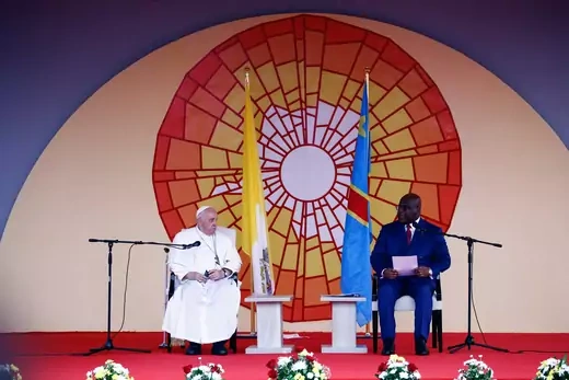 Pope Francis and President of the Democratic Republic of Congo Felix Tshisekedi sit on a stage with a colorful orange, yellow, and white backdrop whilst gazing at one another.