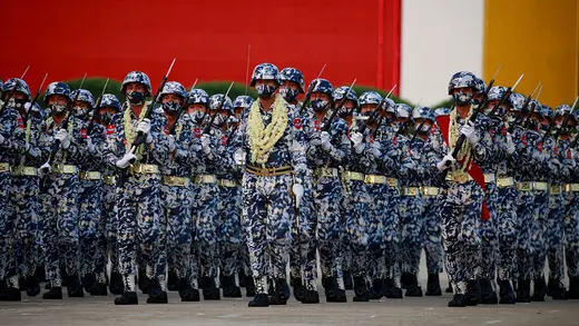 Myanmar soldiers wearing flower garlands as well as blue and white–camouflage fatigues and masks march in formation