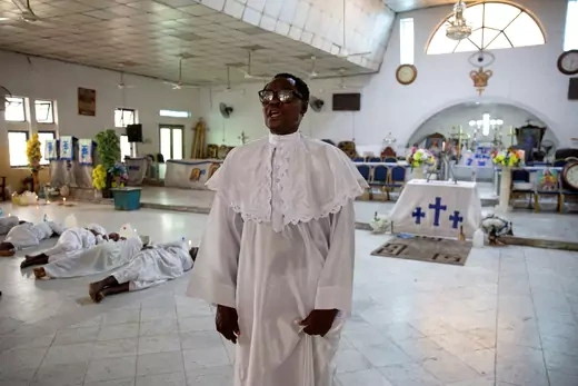 A pastor dressed in white stands before a congregation, some of whom are laying on the ground in prayer.