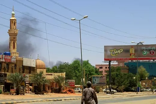 A man walks in the streets of Khartoum, as smoke billows in the sky amidst conflict between rival forces. 