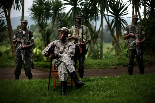 Rebel leader Laurent Nkunda poses outside for a portrait sitting on a chair and surrounded by 3 armed guards.
